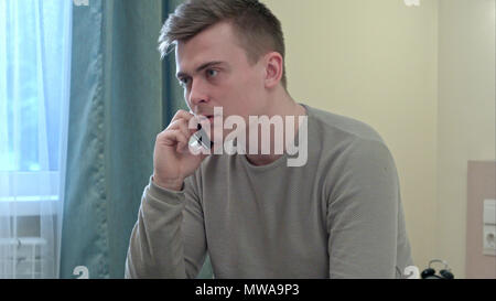 Annoyed young man having a phone call sitting on bed at home Stock Photo