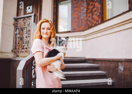 A beautiful young woman with red long hair is holding a small, cute funny big-eyed dog of two flowers, a black-and-white pet of the breed of hichuahua Stock Photo