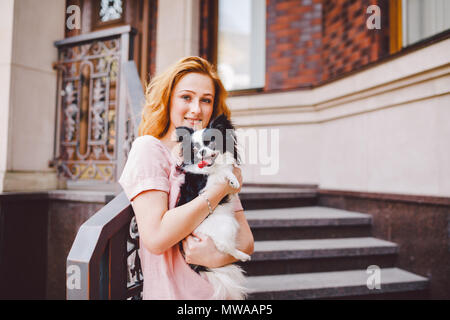 A beautiful young woman with red long hair is holding a small, cute funny big-eyed dog of two flowers, a black-and-white pet of the breed of hichuahua Stock Photo