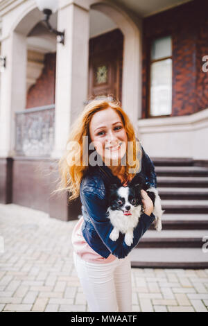 A beautiful young woman with red long hair is holding a small, cute funny big-eyed dog of two flowers, a black-and-white pet of the breed of hichuahua Stock Photo