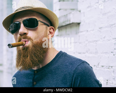 Photo of a bearded man with hat and sunglasses standing outside smoking a cigar. Stock Photo