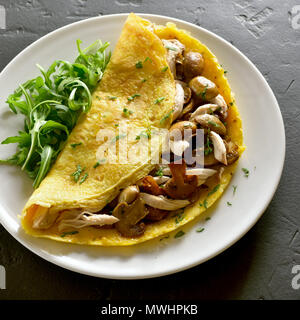 Close up of omelet stuffed with mushrooms, chicken meat, greens on black stone table. Stock Photo