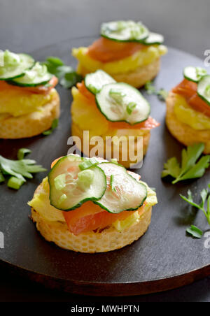 Canape with salmon, scrambled eggs and cucumber slices on black wooden board Stock Photo