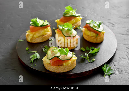 Tasty canape with salmon, scrambled eggs and cucumber slices on black wooden board Stock Photo