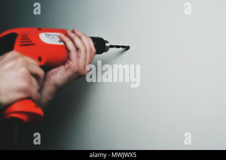 Drilling with a drill in the wall. Man holding a red drill in his hand, the concept of renovation, homemade needlework. Suspending shelves, arranging  Stock Photo