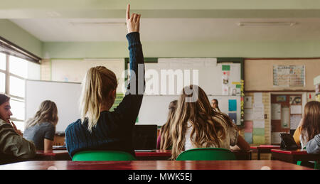 Rear view of female student sitting in the class and raising hand up to ask question during lecture. High school student raises hand and asks lecturer Stock Photo