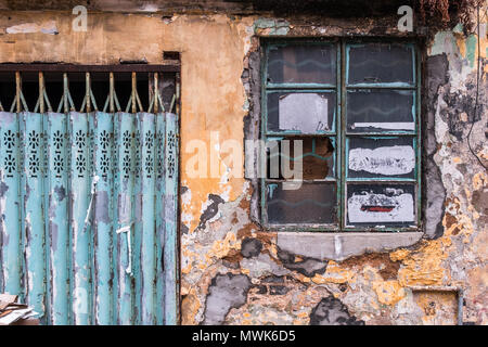 Old rough gray bricks wall with cracked concrete, peeled yellow paint and mold stains. Window with dirty glass in wooden frame. Closed metal door. Stock Photo