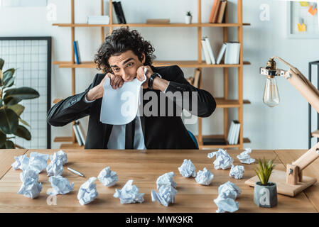 portrait of stressed businessman biting blank paper while while sitting at workplace in office Stock Photo