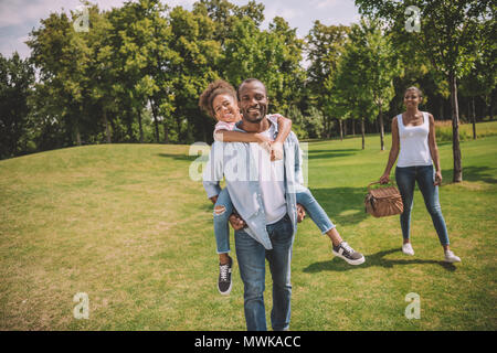 smiling african american young man and little daughter piggybacking together while going on picnic in park Stock Photo