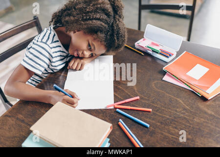 Tired little girl sitting at the table, with her head on her arm, writing in her notebook Stock Photo