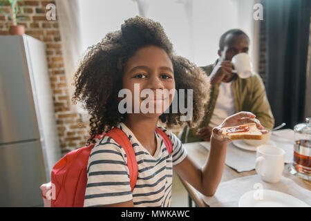 Small girl eating toast with jam at the kitchen table with her dad drinking coffee in the background Stock Photo