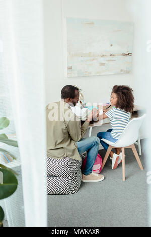 Dad helping his daughter do the homework by explaining the book, while she's sitting at the writing desk with school supplies Stock Photo