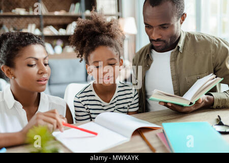Mom and dad helping their little daughter do homework, mother pointing at the notebook and dad holding a textbook Stock Photo