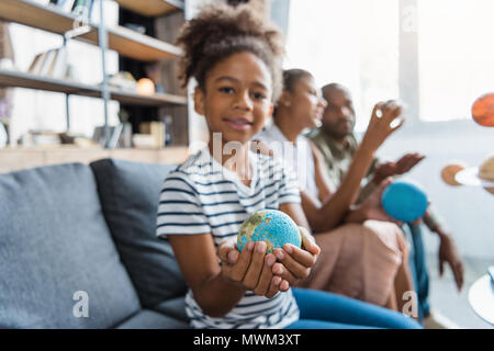 Little girl on a couch with a globe figurine in her hands, while her parents are sitting in the background Stock Photo