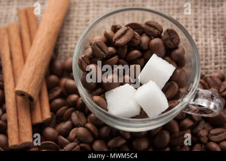 Coffee beans and sugar in glass cup on sackcloth with cinnamon sticks beside. Scattered coffee beans. Stock Photo