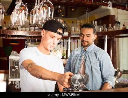 Young trainee bartender pouring beer under supervision from mana Stock Photo
