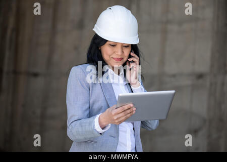 Professional middle aged businesswoman in hard hat talking on smartphone and using digital tablet Stock Photo