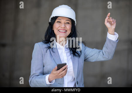 Smiling middle aged asian businesswoman holding smartphone and waving hand Stock Photo
