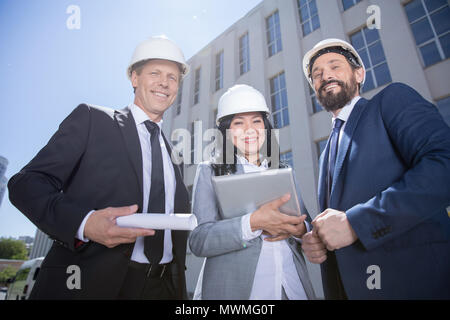 Smiling professional architects in hard hats holding blueprint and using digital tablet Stock Photo