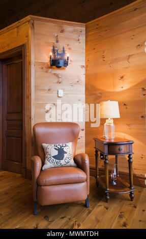 Brown leather sitting chair and wooden side table with lit clear glass lamp in boudoir inside a cottage style flat log profile and timber home Stock Photo