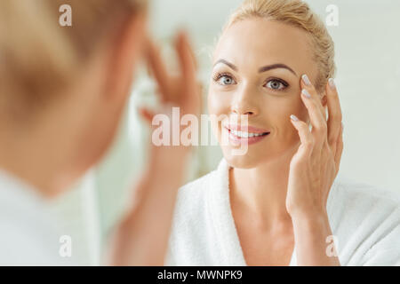 selective focus of beautiful smiling young woman touching face while looking at mirror Stock Photo