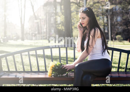 Satisfied girl is calling and looks at flowers. Happy woman is touching by hand the yellow roses and is conducting a conversation on the phone. Women' Stock Photo