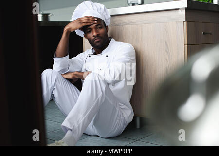 exhausted african american chef sitting on floor at restaurant kitchen and looking away Stock Photo