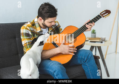 handsome man playing on acoustic guitar and sitting on sofa with dog Stock Photo