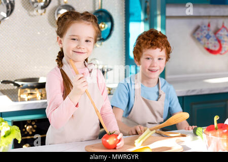adorable happy children in aprons smiling at camera while cooking together in kitchen Stock Photo