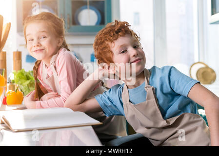 adorable kids in aprons smiling at camera while cooking with cookbook in kitchen Stock Photo