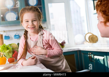 adorable little child in apron smiling at camera while cooking with friend in kitchen Stock Photo