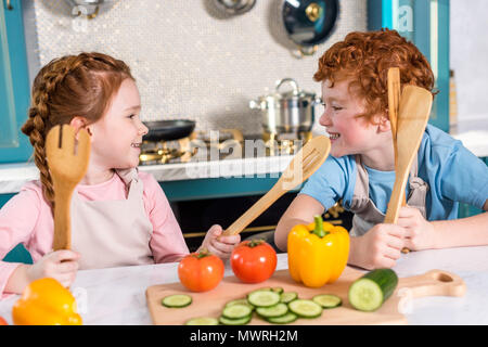 happy kids with wooden utensils smiling each other while cooking together in kitchen Stock Photo