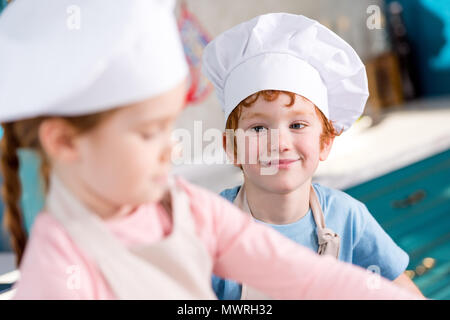 adorable smiling little boy in chef hat looking at friend cooking on foreground Stock Photo