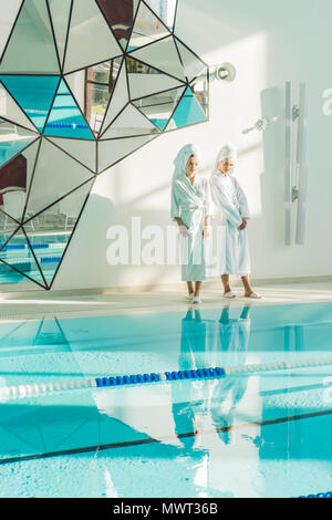 attractive young women in bathrobes standing next to swimming pool at spa center Stock Photo