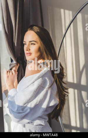 pretty pensive woman in bathrobe standing at window at home Stock Photo