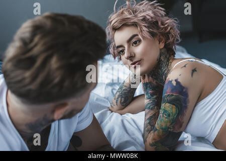 young tattooed couple looking at each other and lying on bed in bedroom Stock Photo