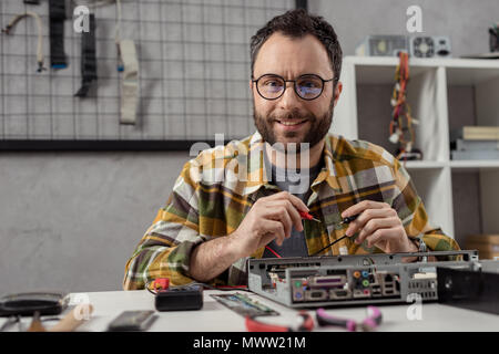 smiling man looking at camera while using multimeter over broken pc Stock Photo