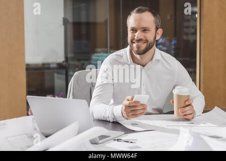 happy architect with paper cup of coffee using smartphone at office Stock Photo