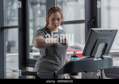 Overweight girl looking at fitness tracker while running on treadmill in gym Stock Photo