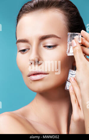 Closeup portrait of beautiful young woman applies the ice to face on a blue background. Skin care concept. Stock Photo