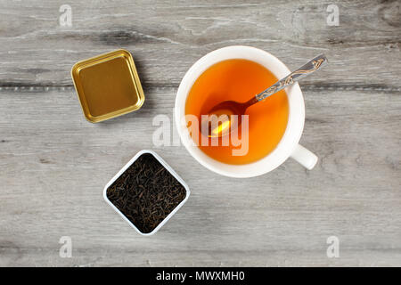 Table top view on cup of hot amber tea with spoon, and caddy full of black dried loose leaves, placed on gray wood desk. Stock Photo
