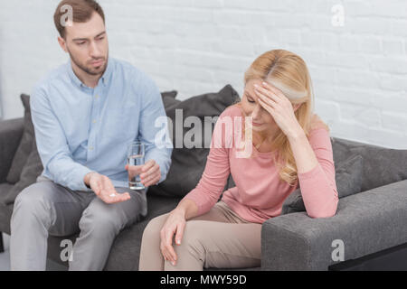 careful son giving glass of water and pills to mother with headache on sofa at home Stock Photo