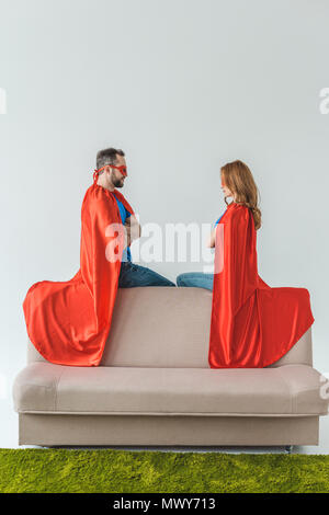 super couple in masks and cloaks sitting on couch and looking at each other on grey Stock Photo