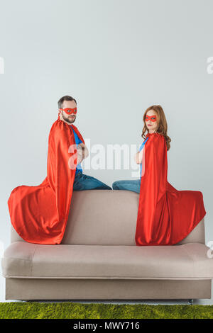 super couple in masks and cloaks sitting on sofa and looking at camera Stock Photo