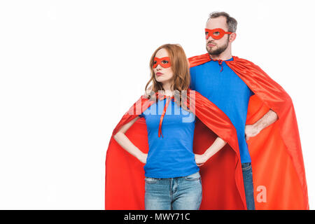 super couple in masks and cloaks standing with hands on waist and looking away isolated on white Stock Photo