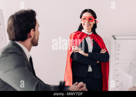 smiling super businesswoman in mask and cape looking at businessman in office Stock Photo