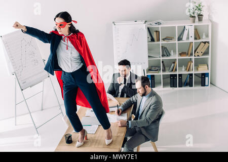super businesswoman in mask and cape standing on table with hand up in office Stock Photo