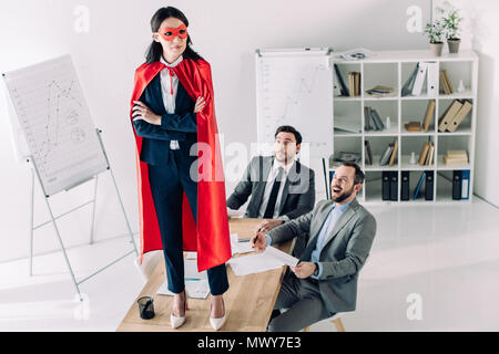 super businesswoman in mask and cape standing on table in office Stock Photo