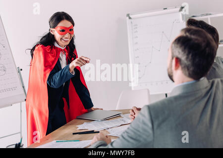smiling super businesswoman in mask and cape pointing on businessmen in office Stock Photo