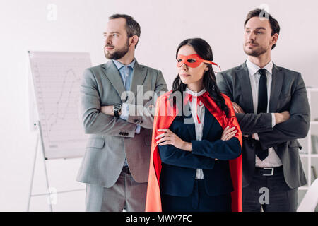 super businesswoman standing with crossed arms in mask and cape with businessmen in office Stock Photo